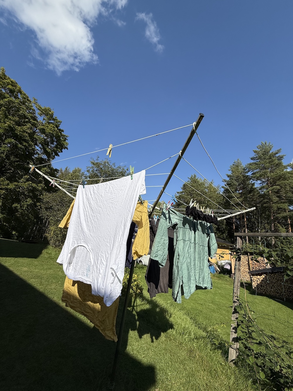 Clothes drying on a rotary washing line in a garden on a sunny day. White t-shirt, yellow and green shirts, and dark items hanging against a bright blue sky with forest trees surrounding the yard