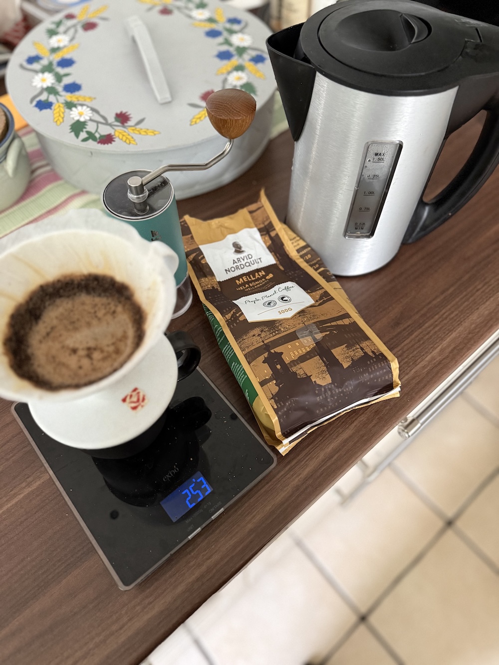 Coffee brewing setup on a wooden surface showing a Commandante hand grinder with wooden knob, a white V60 dripper with filter paper on a mug, a bag of Arvid Nordquist Mellanrost coffee, a digital scale reading 253g, and a stainless steel electric kettle.