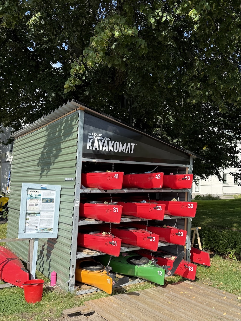 A green wooden shed labeled 'KAYAKOMAT' with numbered red, yellow, and green kayaks stored horizontally on racks, functioning as a self-service kayak rental station