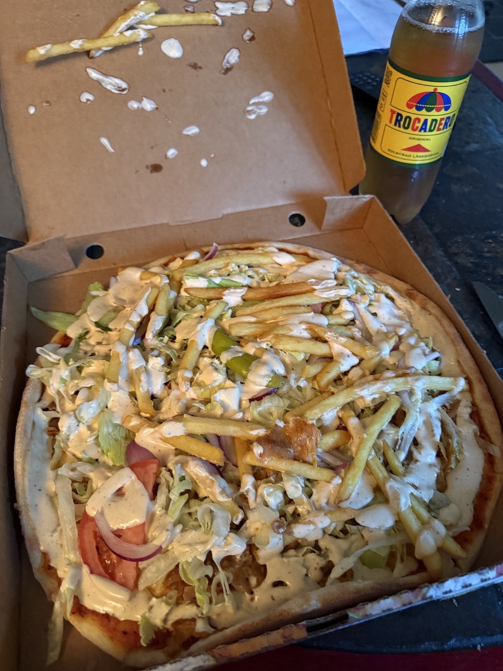 A pizza in a delivery box topped with french fries, lettuce, tomatoes, onions, and kebab meat with white garlic sauce drizzled on top. A bottle of Trocadero soda sits beside it.